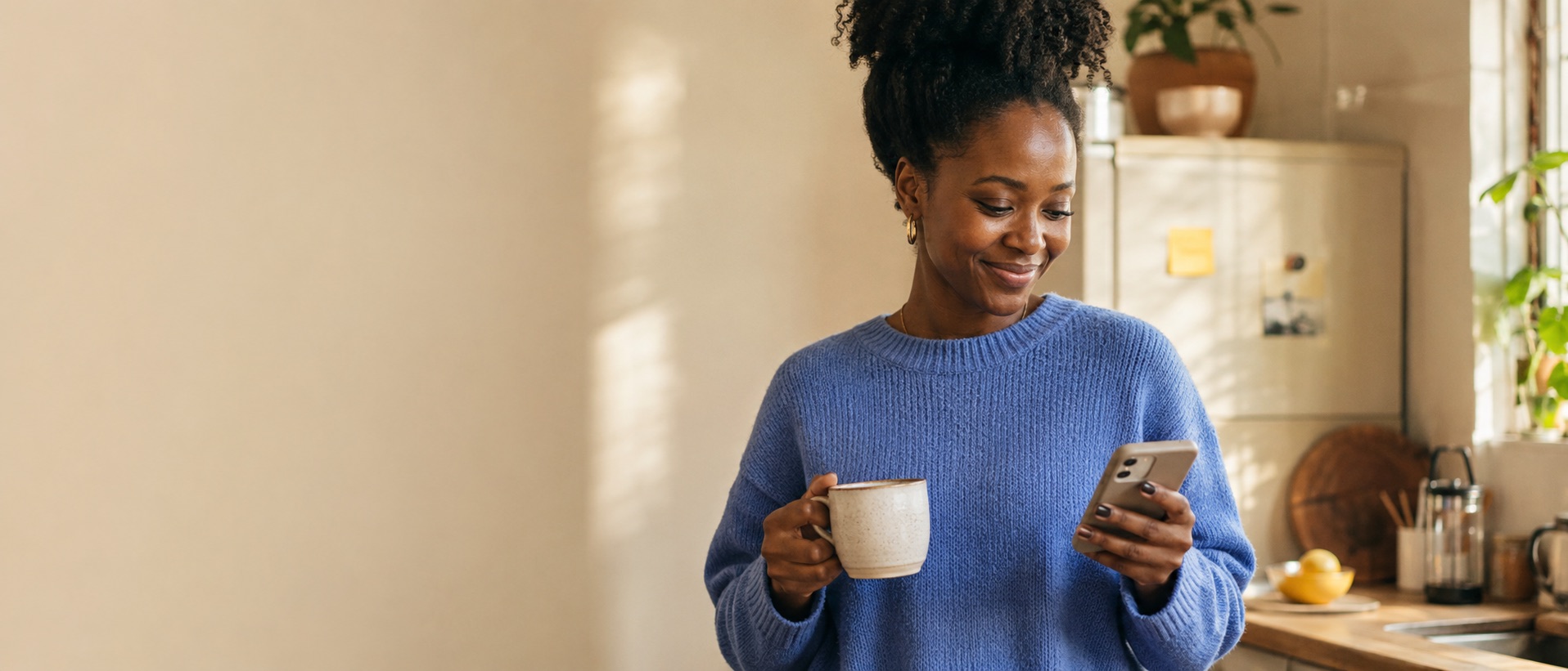 A woman smiles at a well-timed message on her phone, coffee in hand, in her sunlit kitchen.